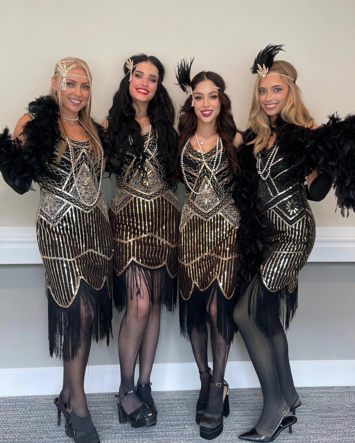 Four women posing in black and gold sequined flapper dresses with fringe and feathers.