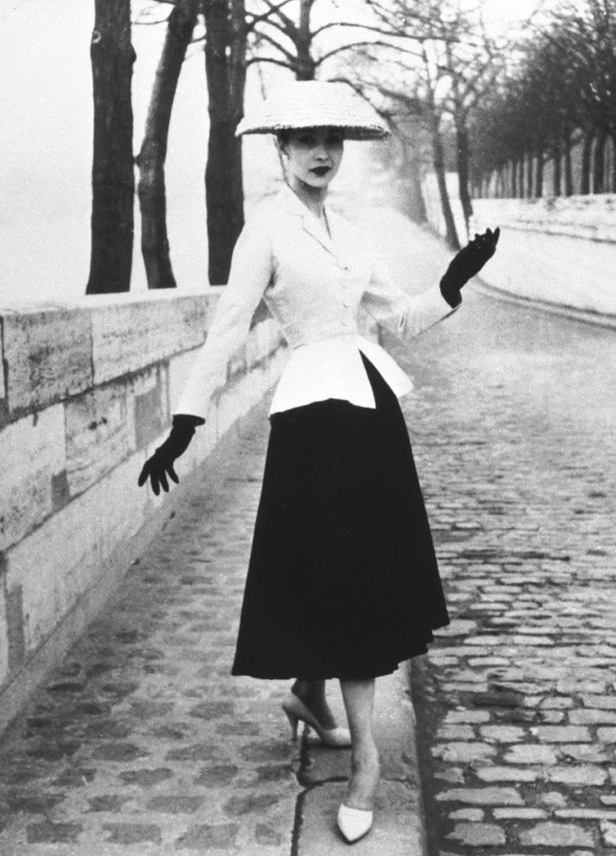 Woman wearing a cinched waist jacket, wide-brimmed hat, and voluminous skirt walking on cobblestones.