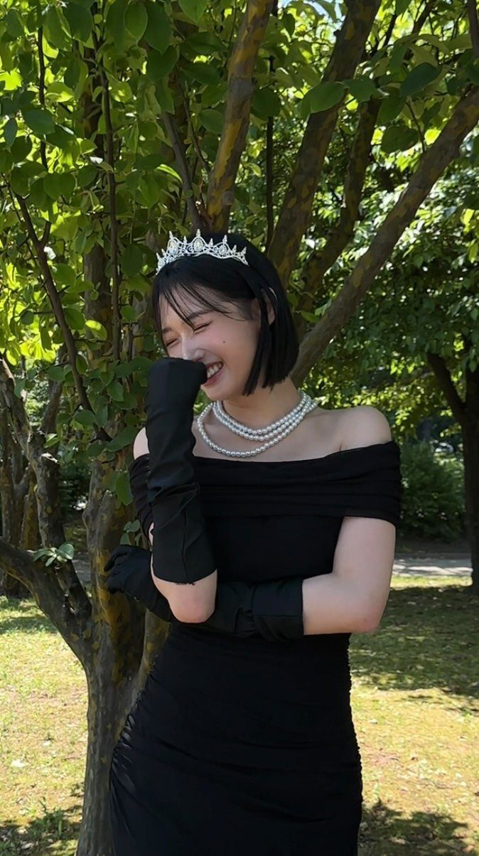 Woman in black off-the-shoulder dress, long gloves, pearls, and a tiara standing by trees.
