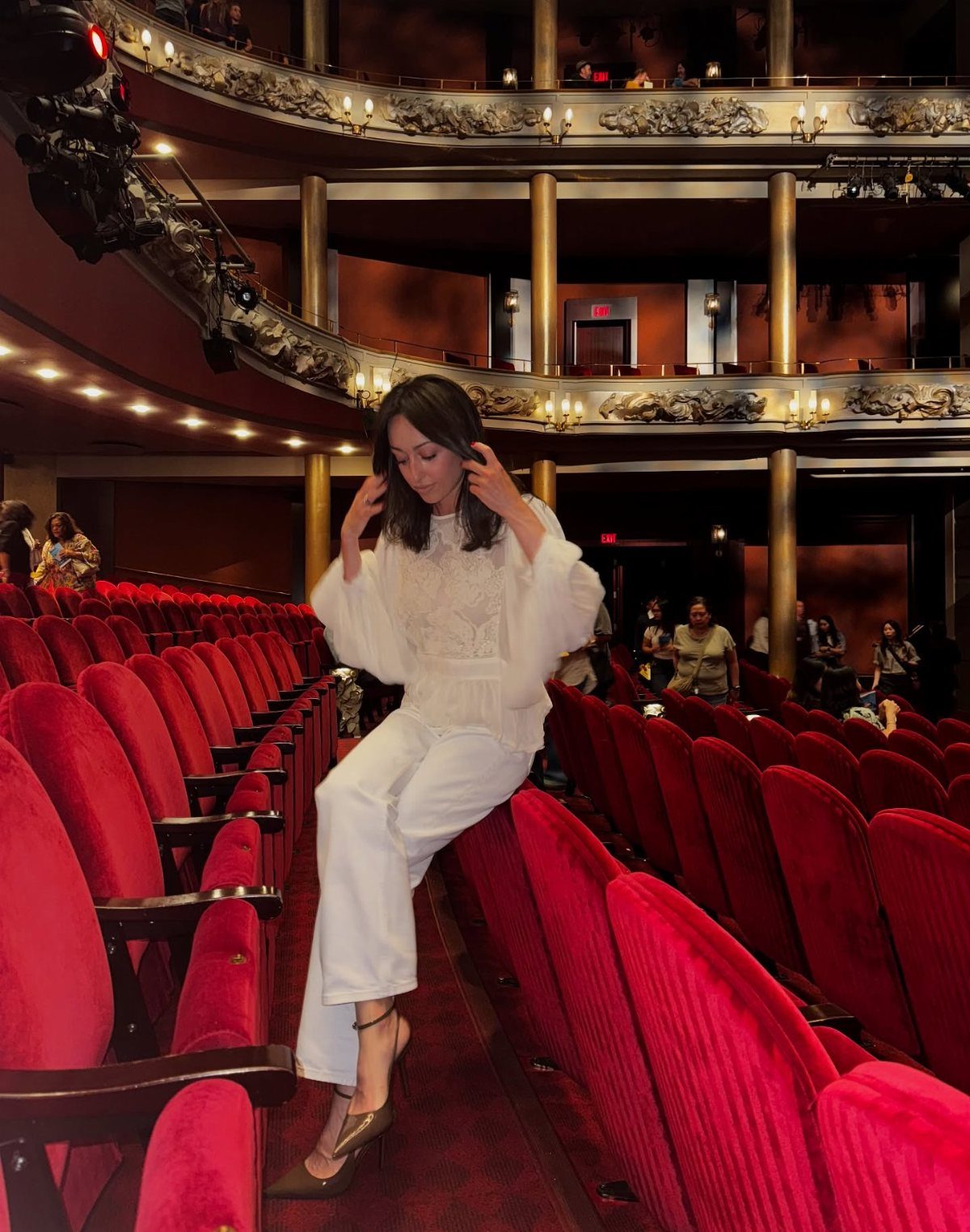 Woman in white wide-leg trousers and blouse sitting in red theater seats.