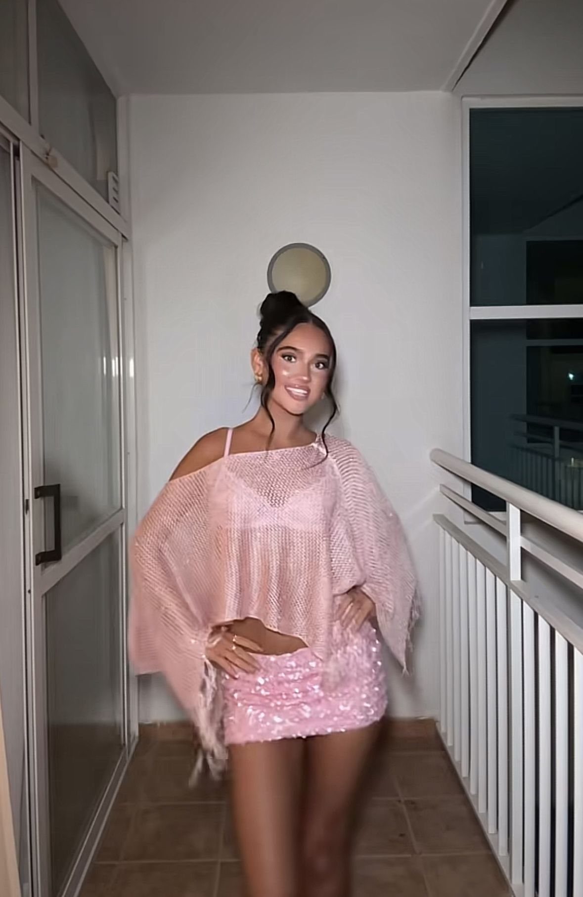 Young woman posing in a pink sequined mini skirt and knit off-the-shoulder top on a balcony.