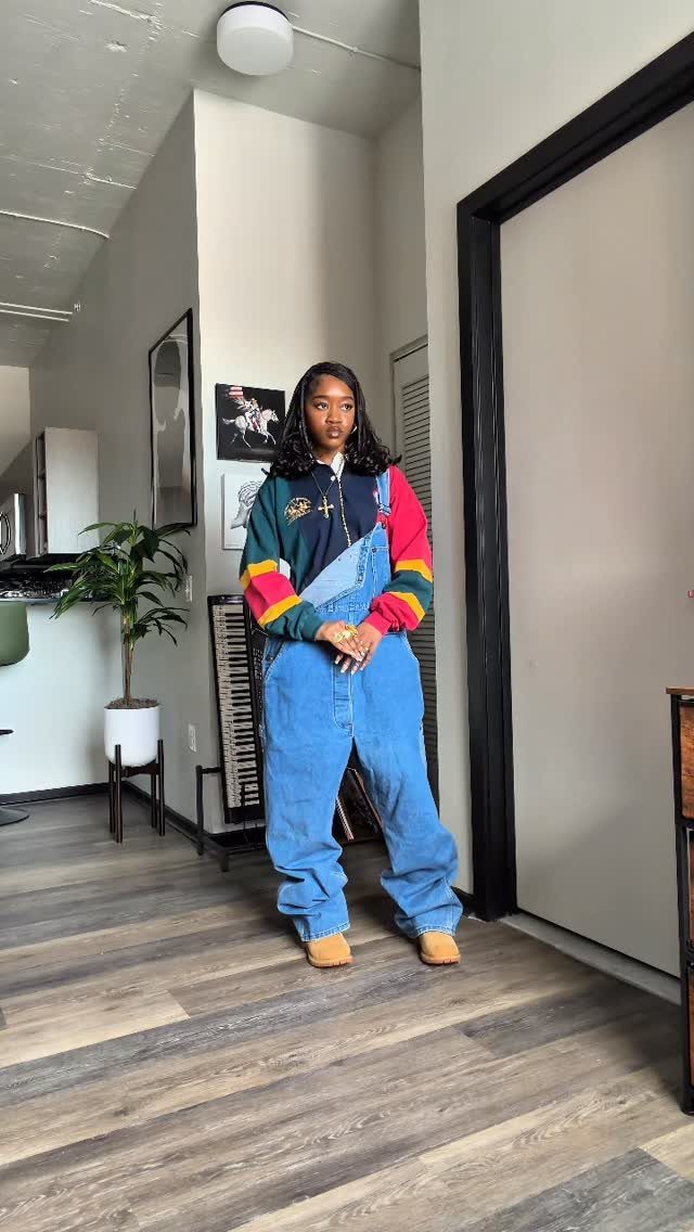 Woman wearing baggy denim overalls and a colorful striped shirt standing in a room.