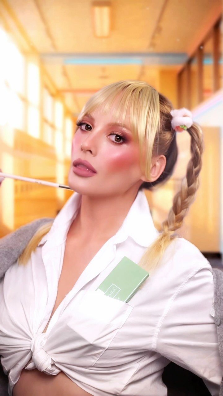 A blonde woman with pigtails wearing a white tied-up shirt in a warm hallway.