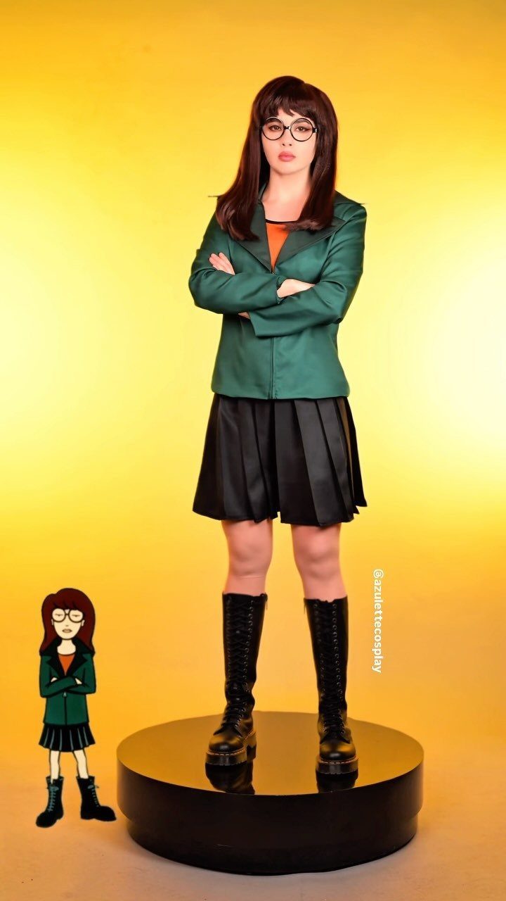 Woman in a green blazer, black pleated skirt, and combat boots posing against a yellow background.