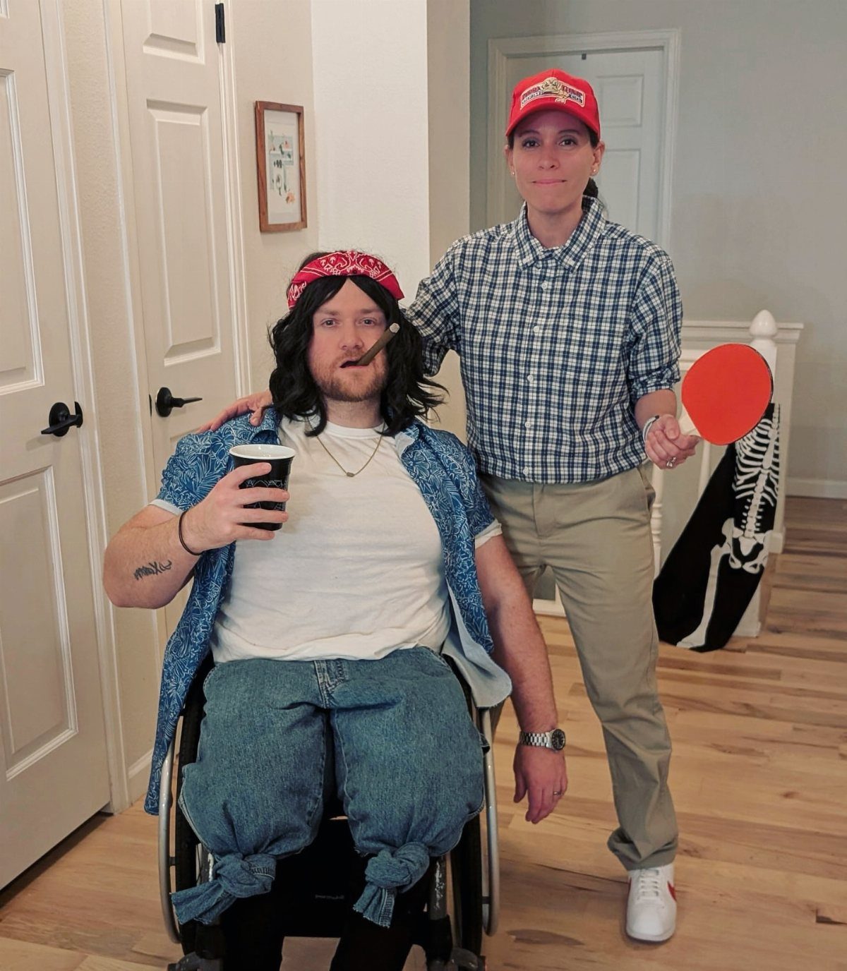 Two people dressed in costumes, one sitting in a wheelchair and one standing with a ping pong paddle.