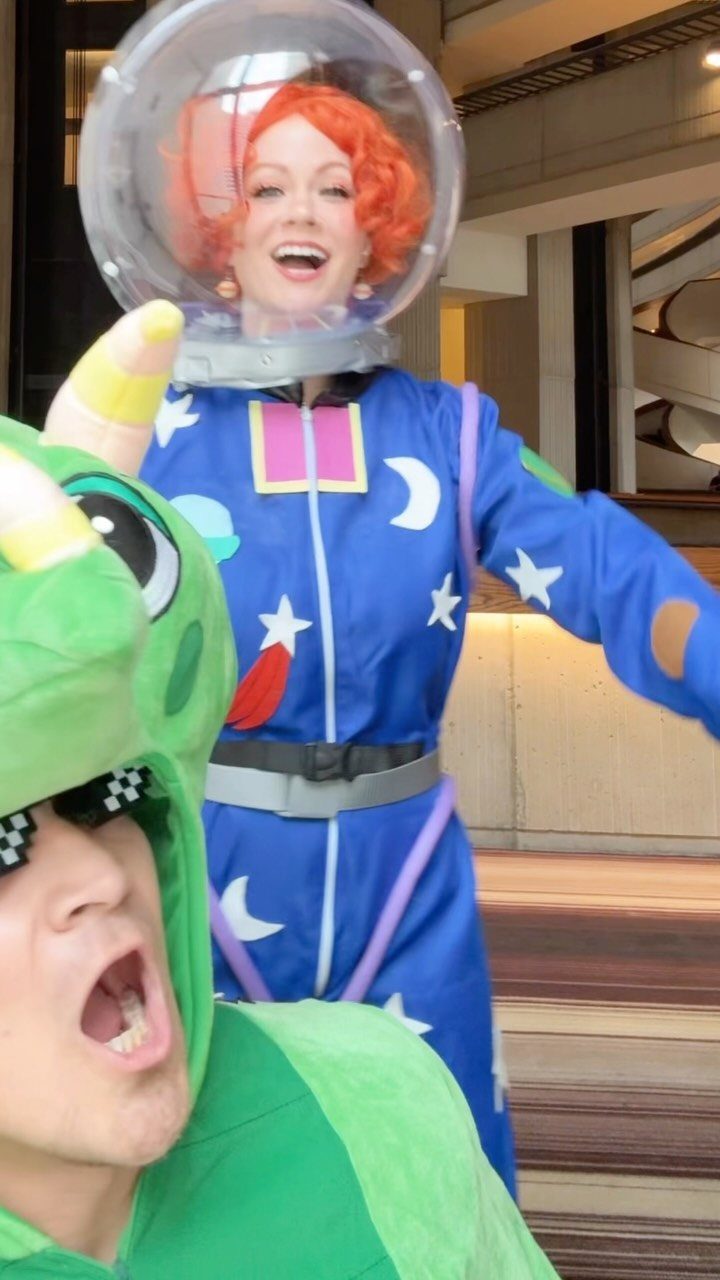 Woman with orange hair in a blue star-patterned jumpsuit and bubble helmet smiling.