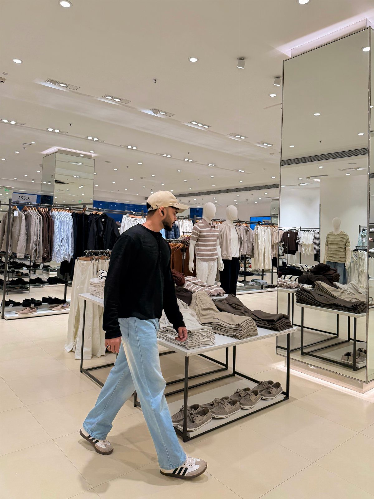 Man in black shirt and light blue jeans walking through a clothing store wearing Adidas sneakers.