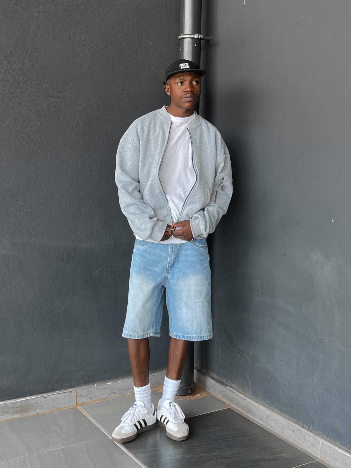 Man wearing a grey bomber jacket, denim shorts, and white Adidas Samba sneakers.