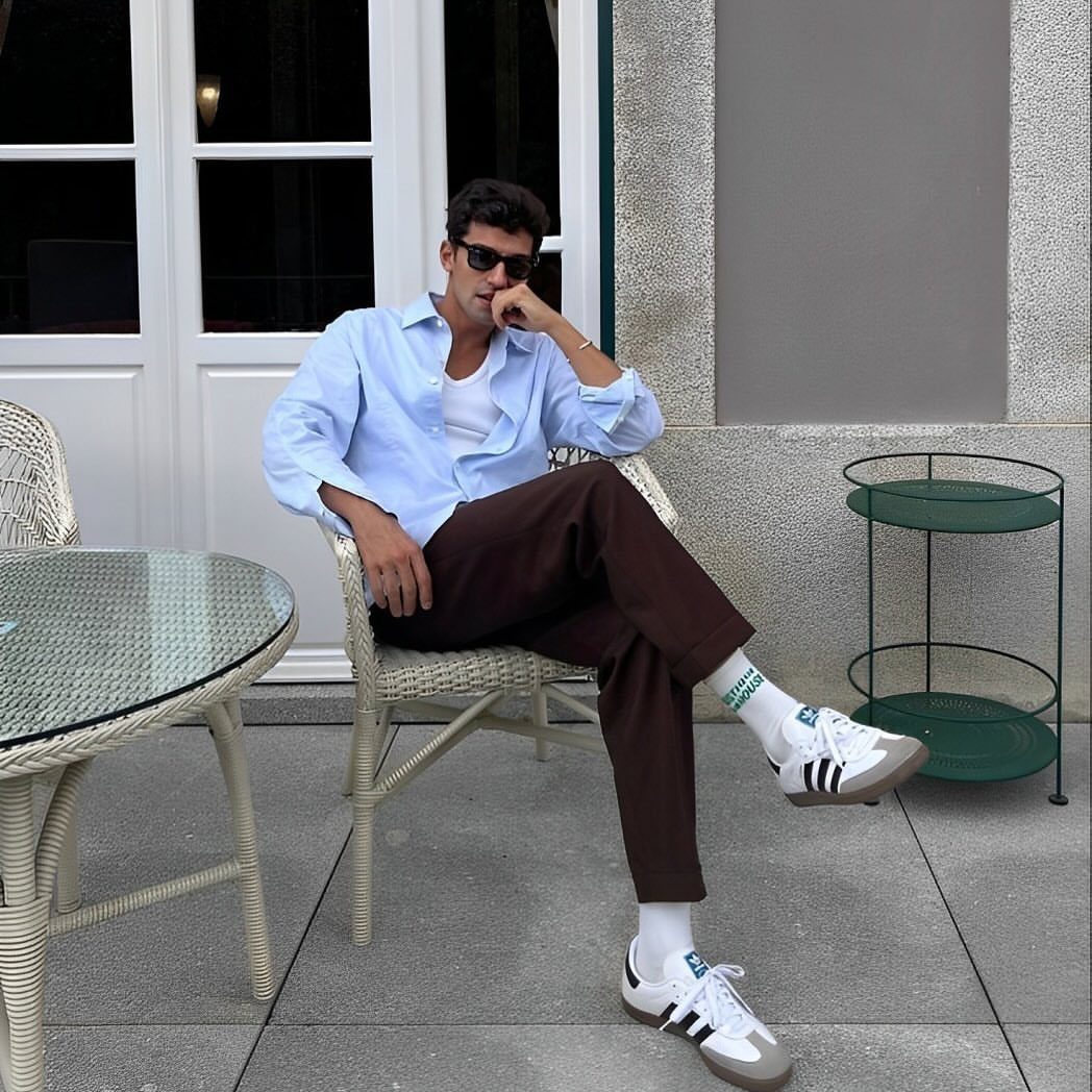 Man in light blue shirt and brown trousers wearing Adidas Samba sneakers while sitting outdoors.