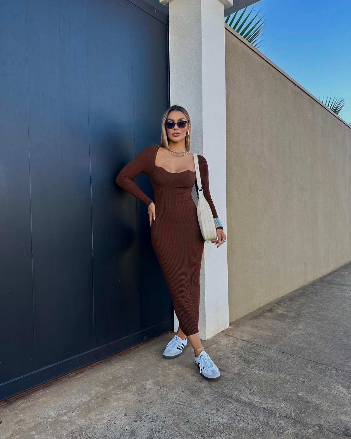 Woman in a brown ribbed midi dress and white sneakers posing against a blue wall.