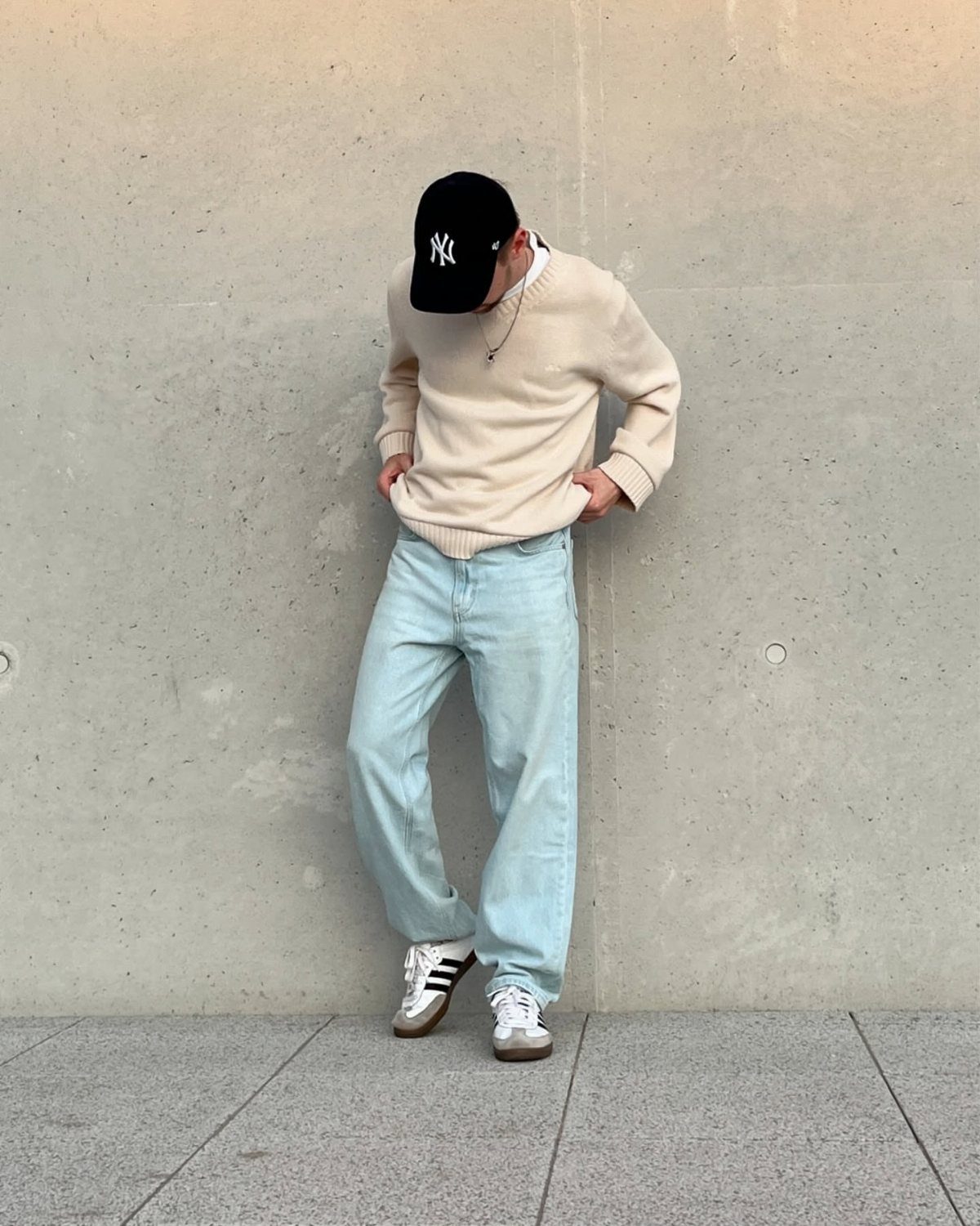 A person in light blue jeans, a cream sweater, and Adidas Samba sneakers leaning against a wall.