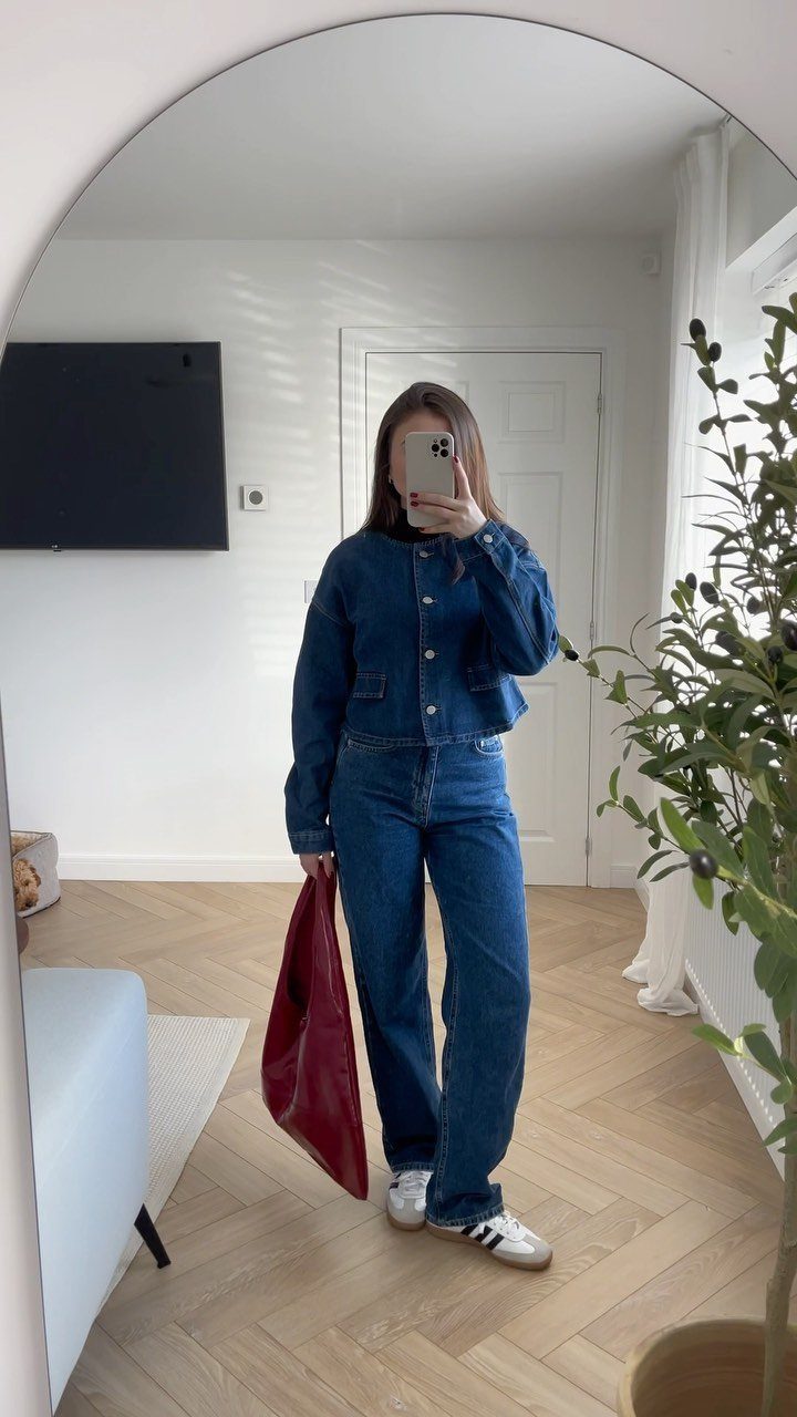 A person posing in a denim jacket, blue jeans, and white sneakers.