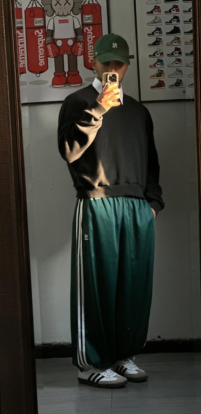 A person posing in a black sweatshirt, green Adidas track pants, and white Adidas Samba sneakers.