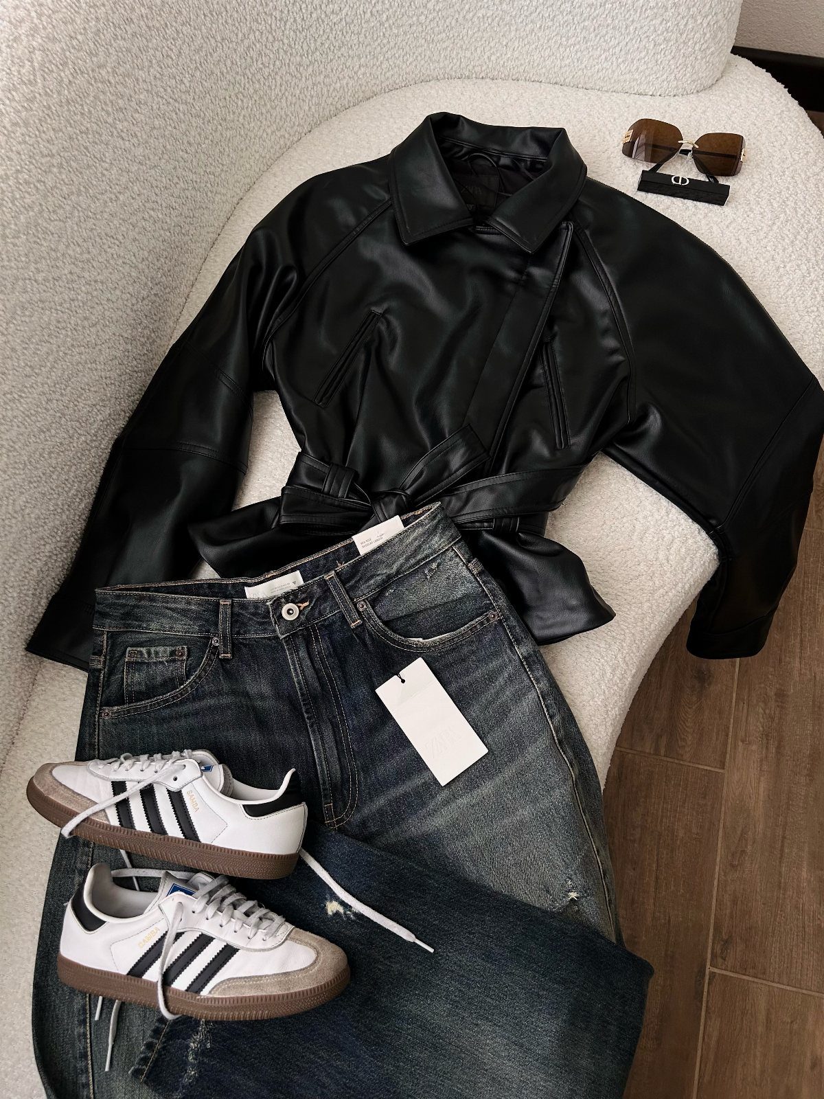 Black leather jacket, blue denim jeans, and white Adidas Samba sneakers on a white chair.