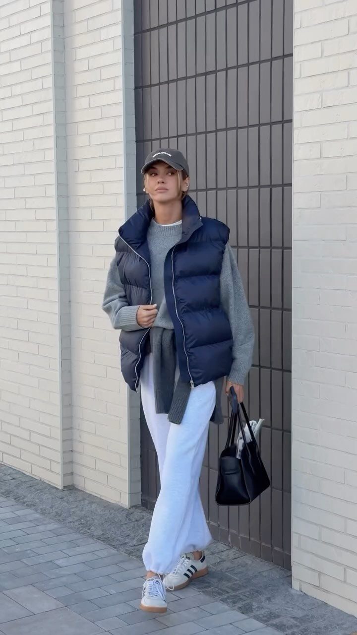 Woman wearing a navy puffer vest, grey sweater, white pants, and Adidas Samba sneakers walking outdoors.