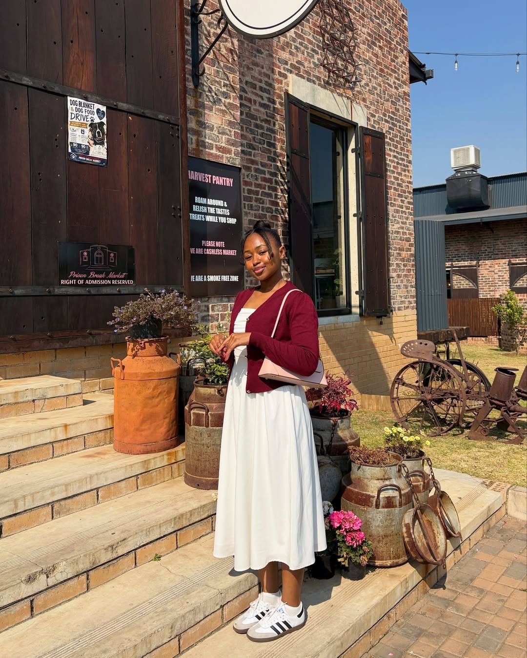 Woman in a white midi dress, maroon cardigan, and white Adidas Samba sneakers standing on steps.