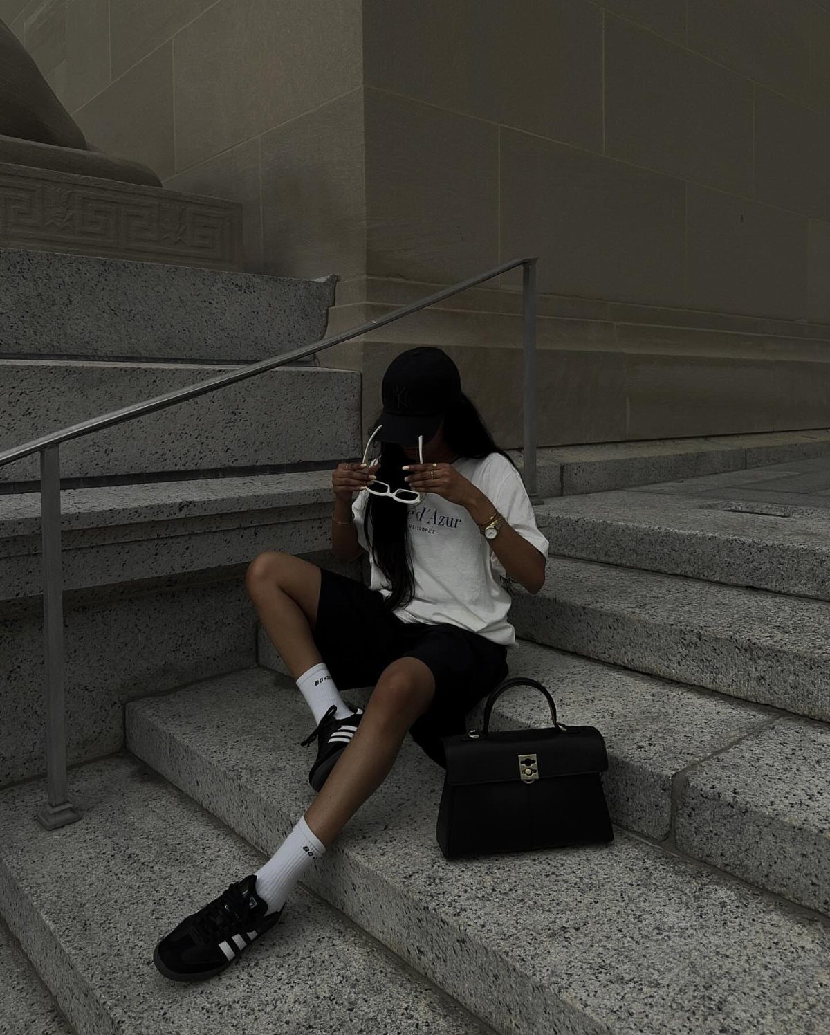 Person sitting on stone stairs wearing a white t-shirt, black shorts, and Adidas Samba sneakers.