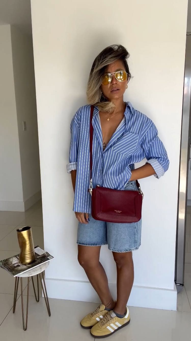 A woman posing in a striped shirt, denim shorts, and yellow Adidas Samba sneakers.