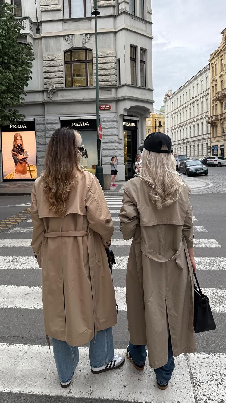 Two women wearing tan trench coats and blue jeans walking across a city street.