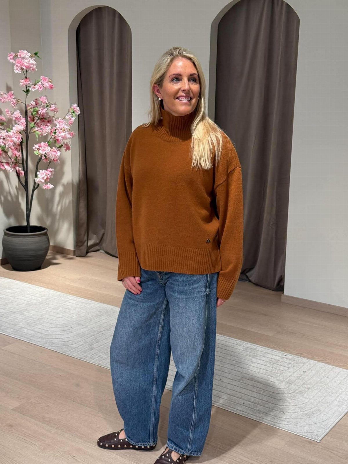 Blonde woman wearing a brown turtleneck sweater and blue barrel jeans standing indoors.
