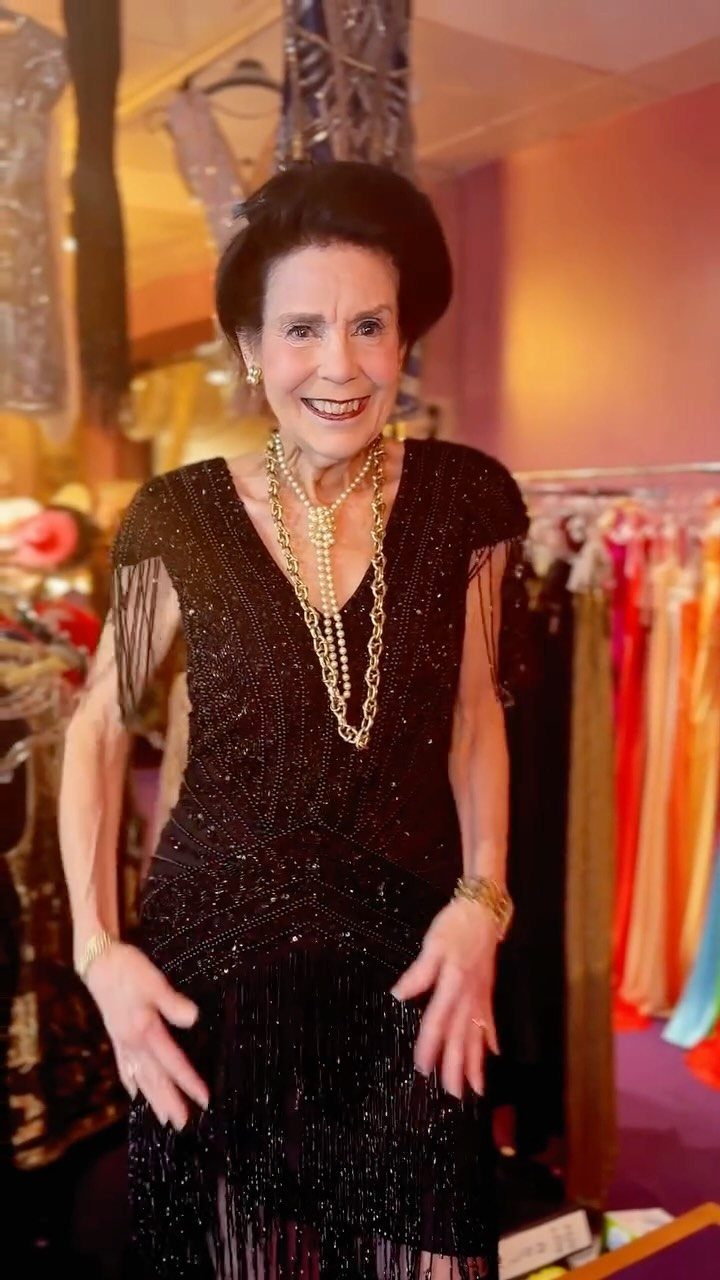 An older woman smiling in a black beaded cocktail dress with fringe details and gold jewelry.