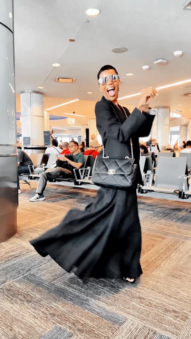 A person posing in an airport wearing an all-black outfit with a long skirt and blazer.