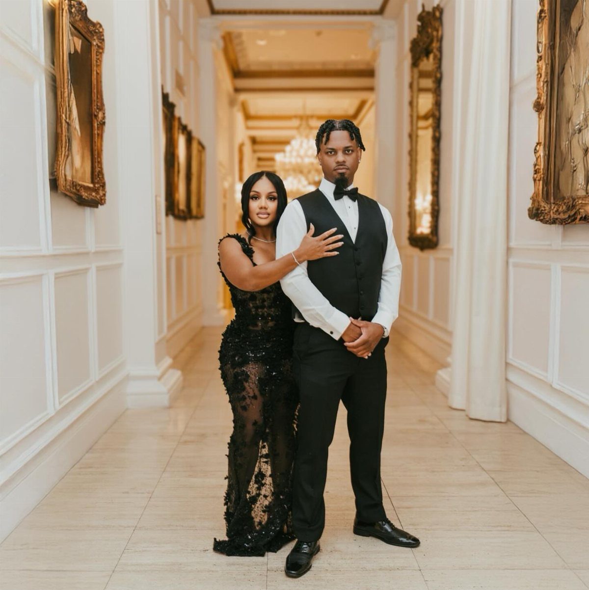 A woman in a black lace dress and a man in a tuxedo posing in a hallway.