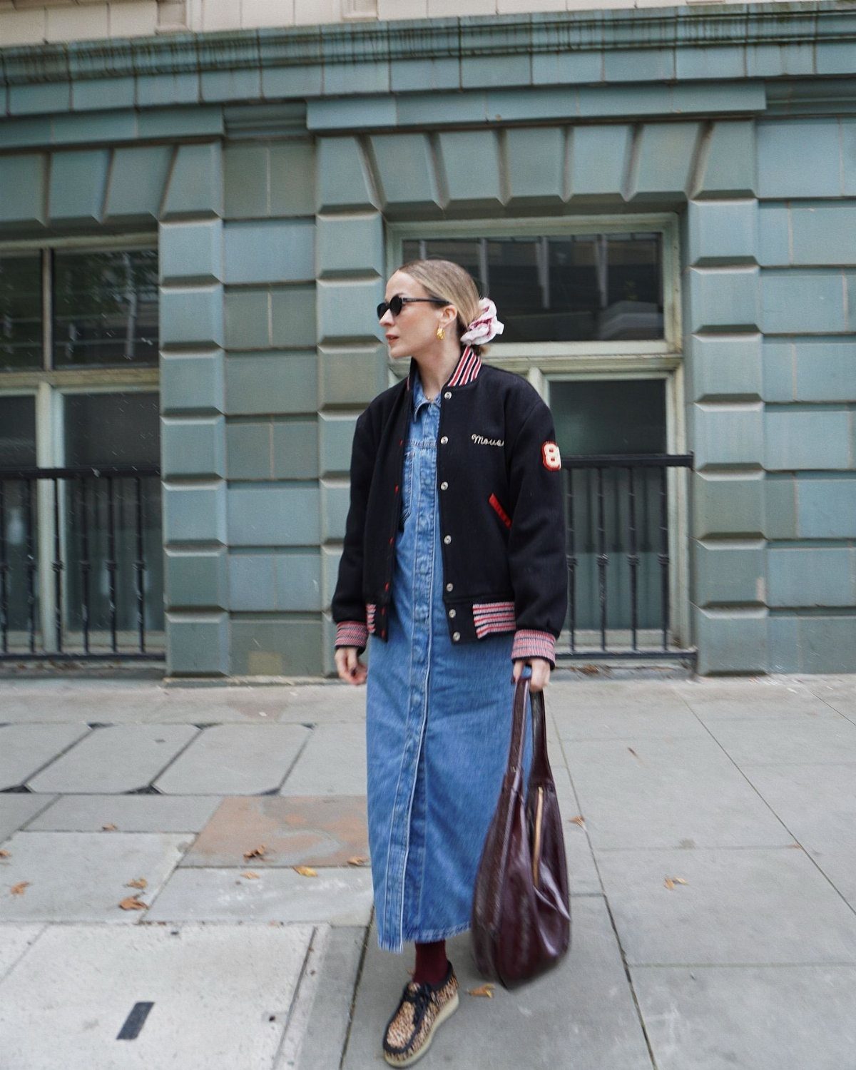 Woman wearing a long denim dress, black bomber jacket, dark socks, and loafers on a city street.