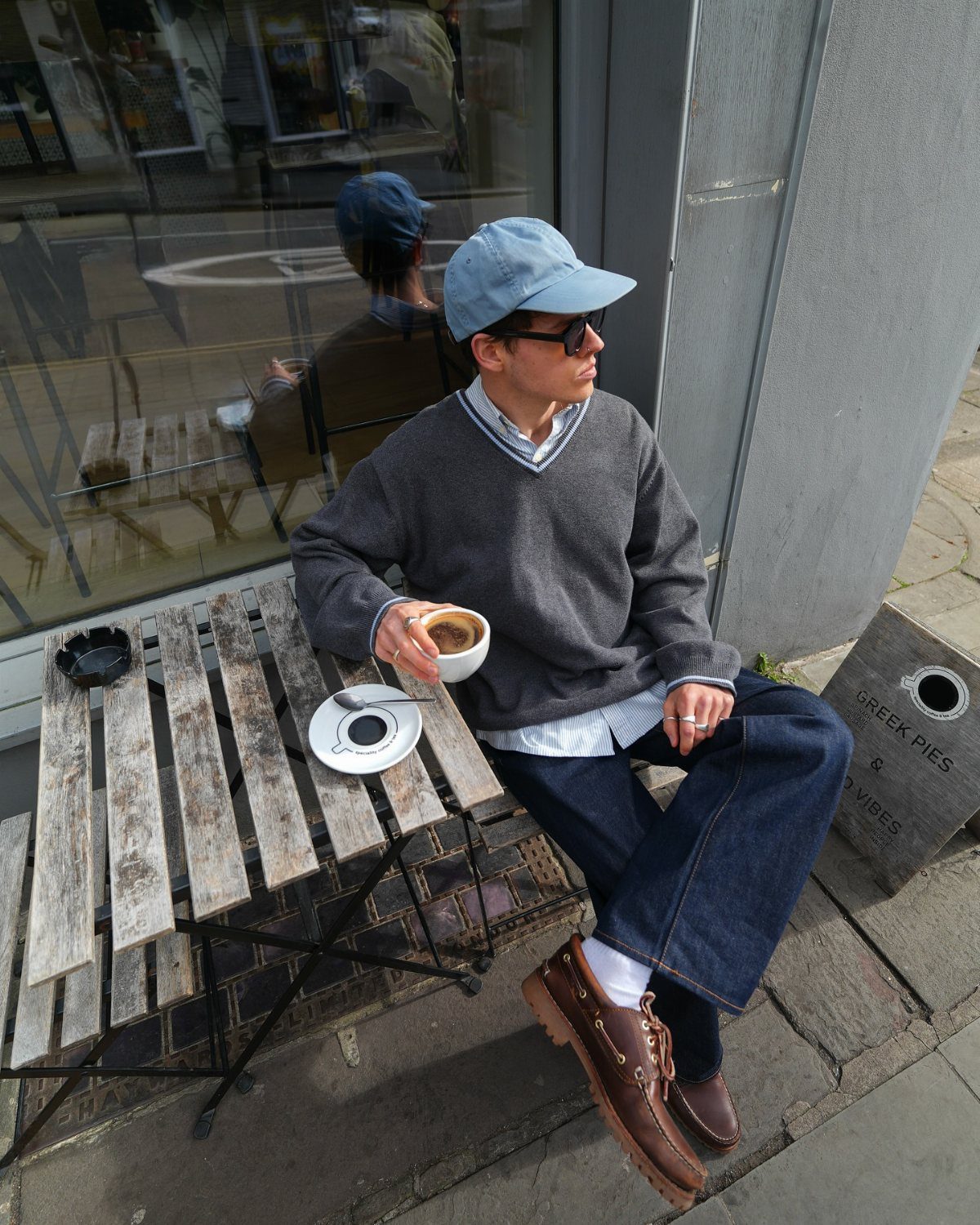 Person wearing a grey sweater, dark denim jeans, and brown boat shoes sitting at a cafe table.