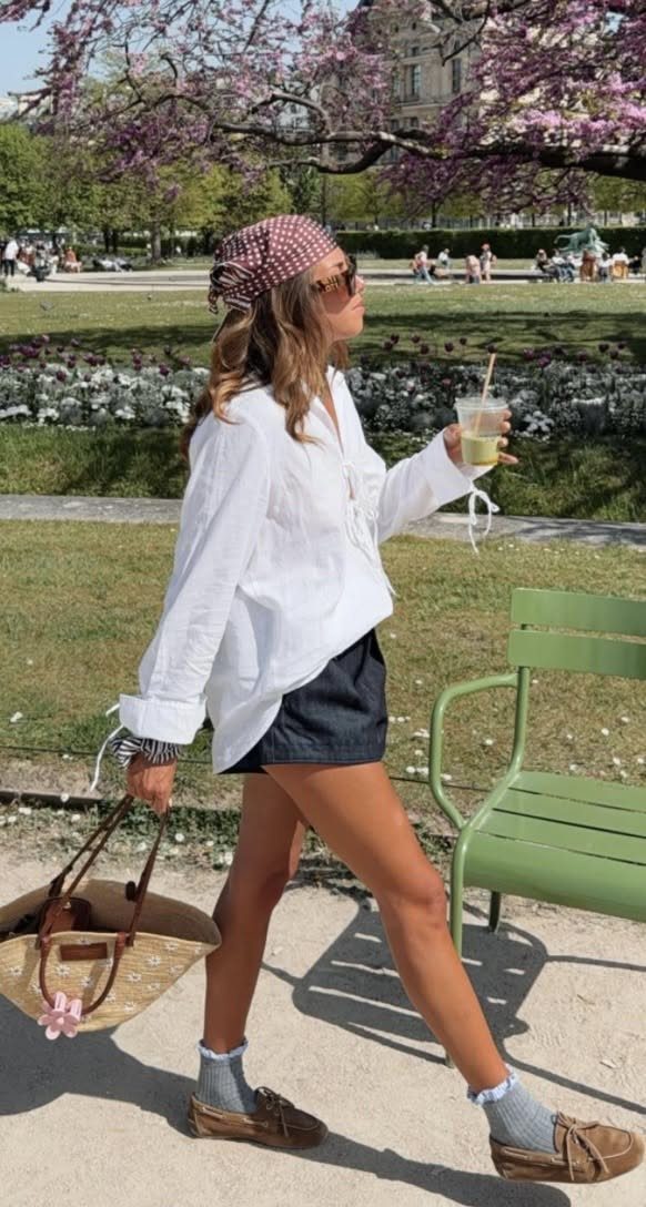 Woman walking through a sunny park wearing a white linen shirt, dark shorts, and brown boat shoes.