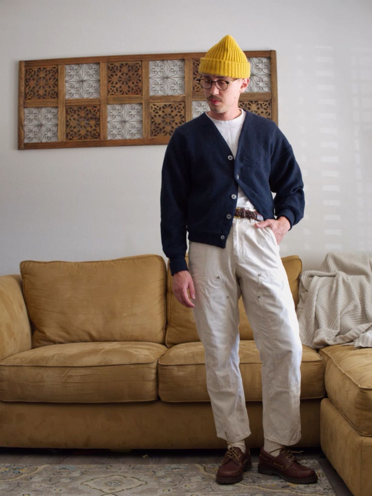 Man in a navy cardigan, cream trousers, and brown boat shoes standing by a sofa.
