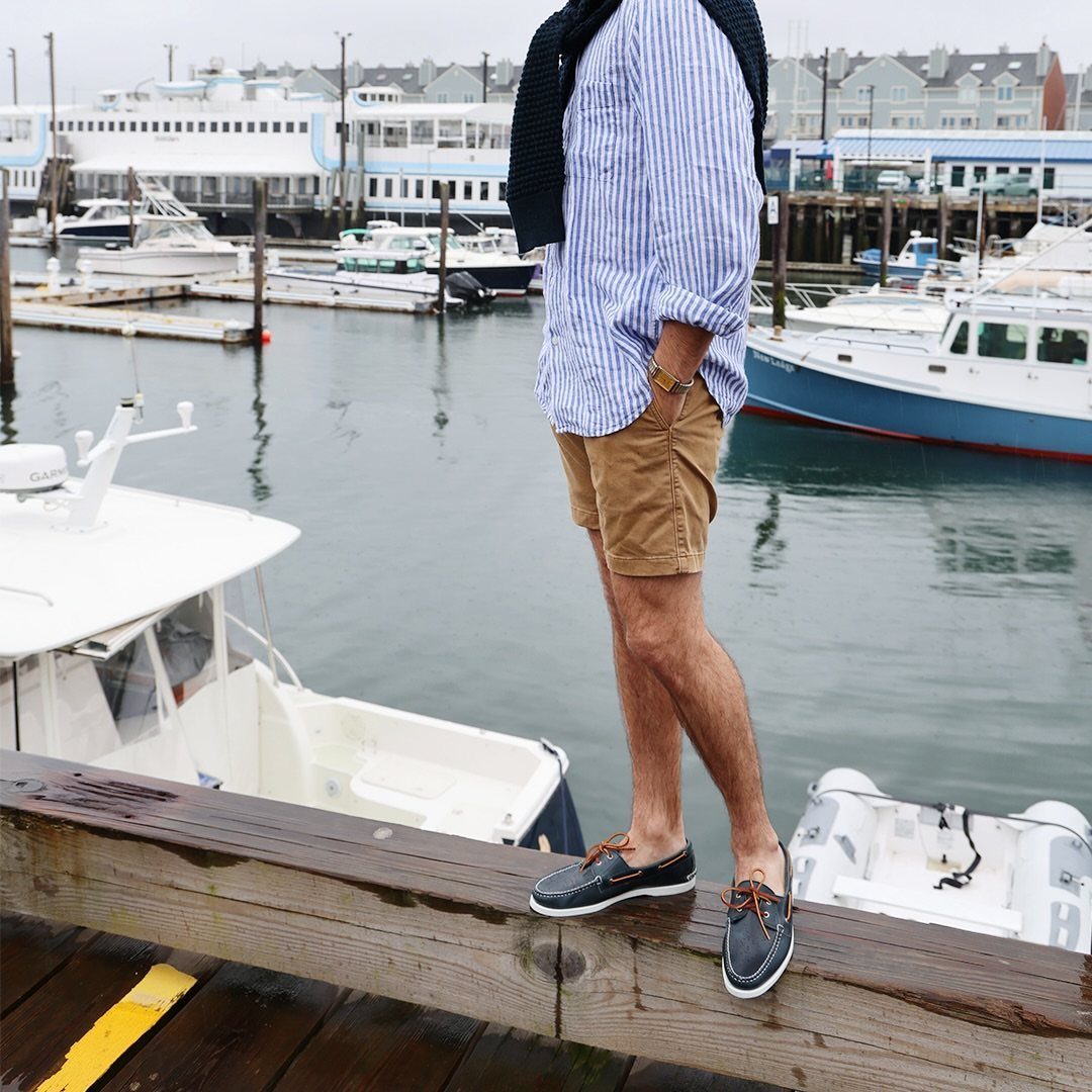 Man in a striped shirt and tan shorts standing on a wooden pier by boats.