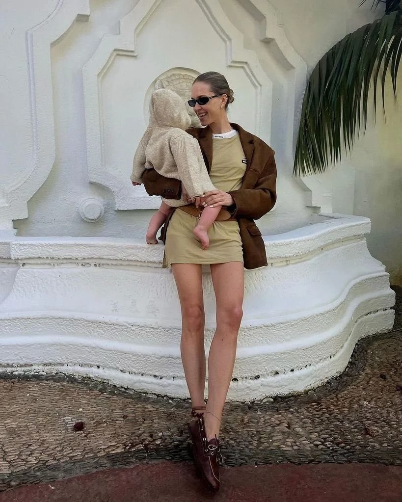 Woman in a tan dress and brown blazer holding a baby in front of a white wall.