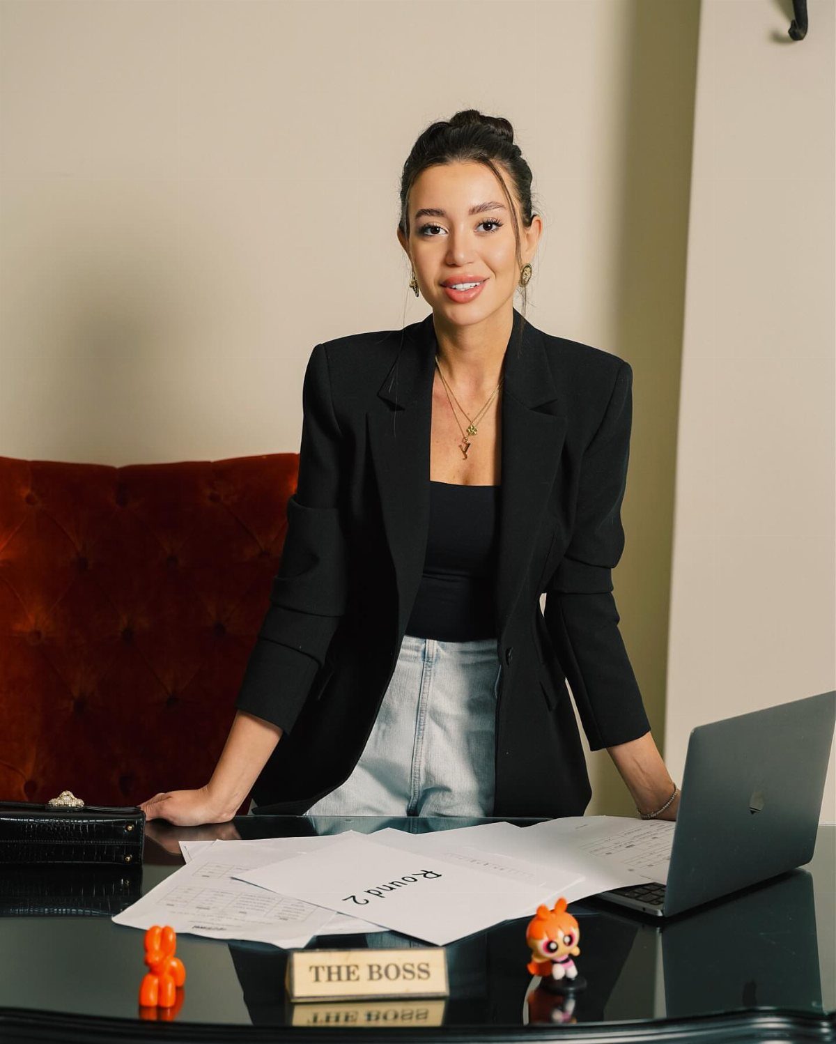 Woman in a black blazer and light blue jeans standing behind a desk with a laptop.