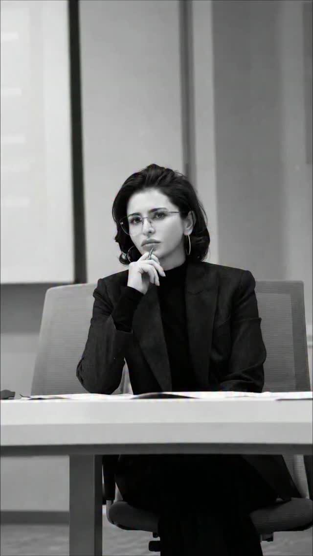 A woman in a professional blazer and turtleneck sitting at an office desk.