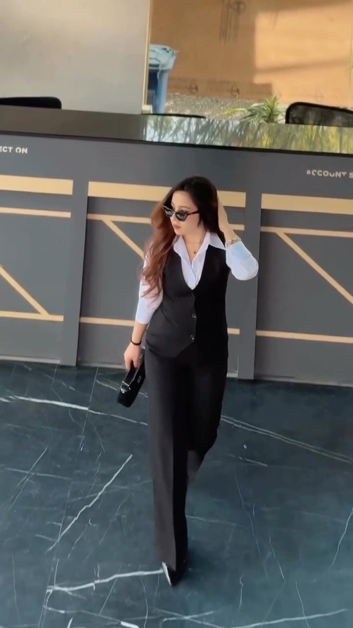A woman in a black waistcoat and trousers walking while wearing sunglasses and carrying a clutch.