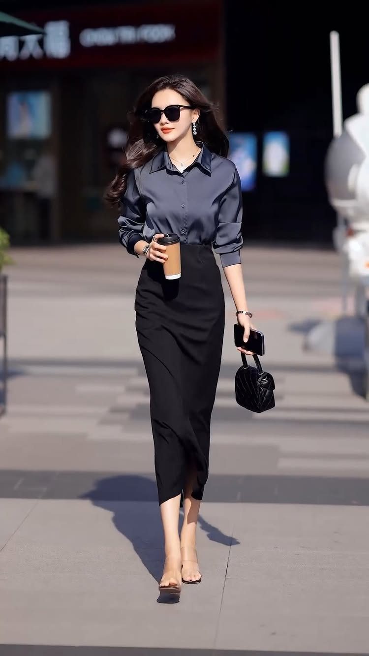 A woman in professional attire walking through a city street while carrying coffee and a handbag.