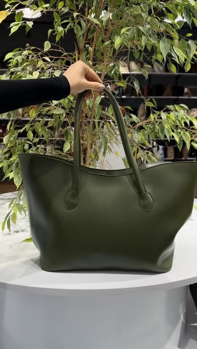 A hand holding the handle of a large olive green leather tote bag on a white table.