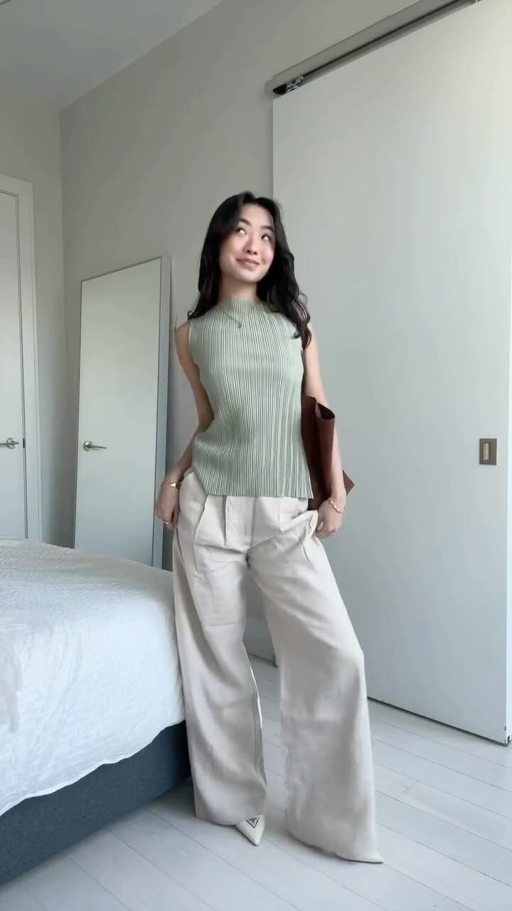 A woman posing in a green ribbed top and beige wide-leg trousers in a bright room.