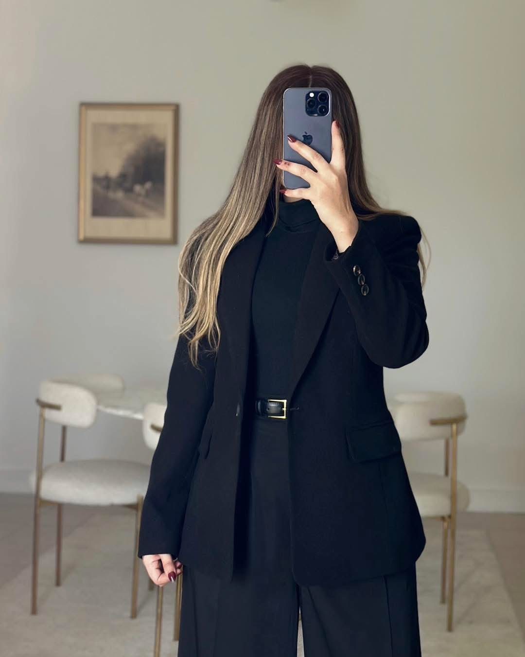 Woman in a black blazer and turtleneck taking a mirror selfie in a bright room.