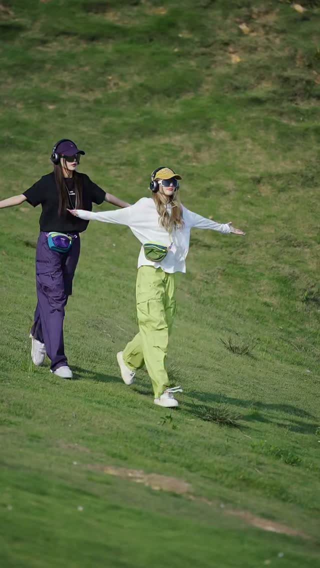 Two women wearing cargo pants and sporty accessories walking across a green grassy hill.