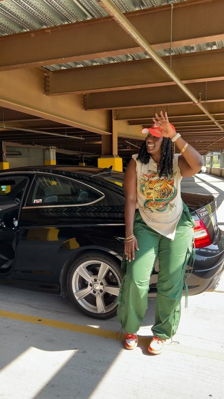 A woman in green cargo pants and a graphic T-shirt posing by a black car.