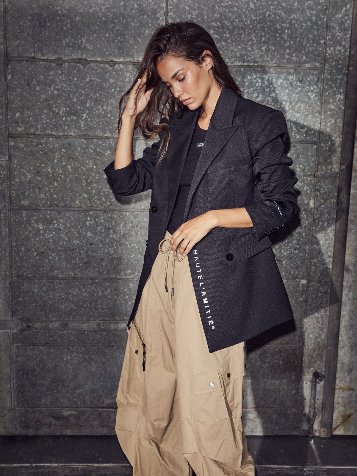 Woman wearing a dark blazer, black top, and tan cargo pants against a stone wall.