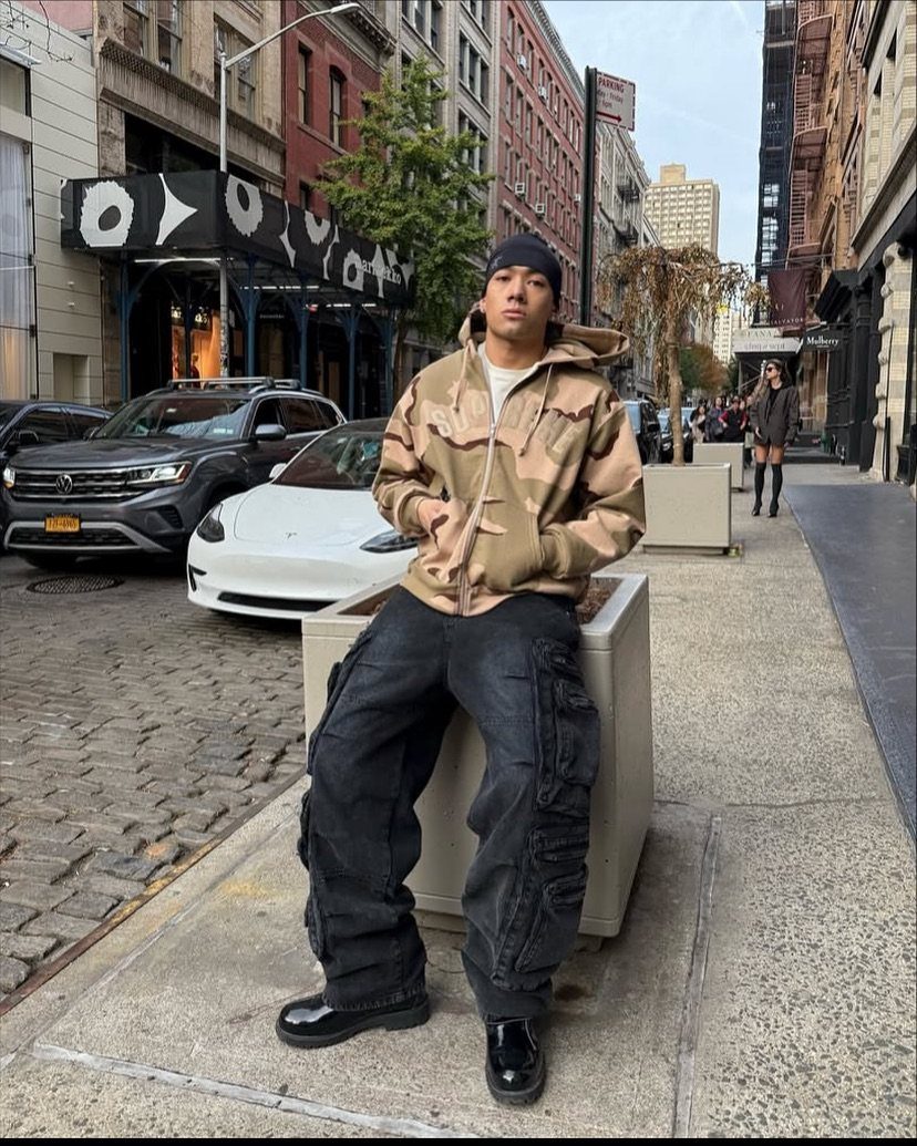 Man sitting on a city planter wearing black cargo pants and a tan oversized hooded jacket.