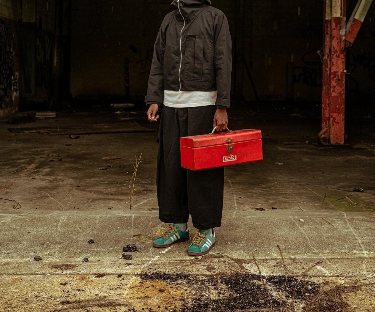 Person wearing black cargo pants and a dark jacket holding a red toolbox in an industrial setting.