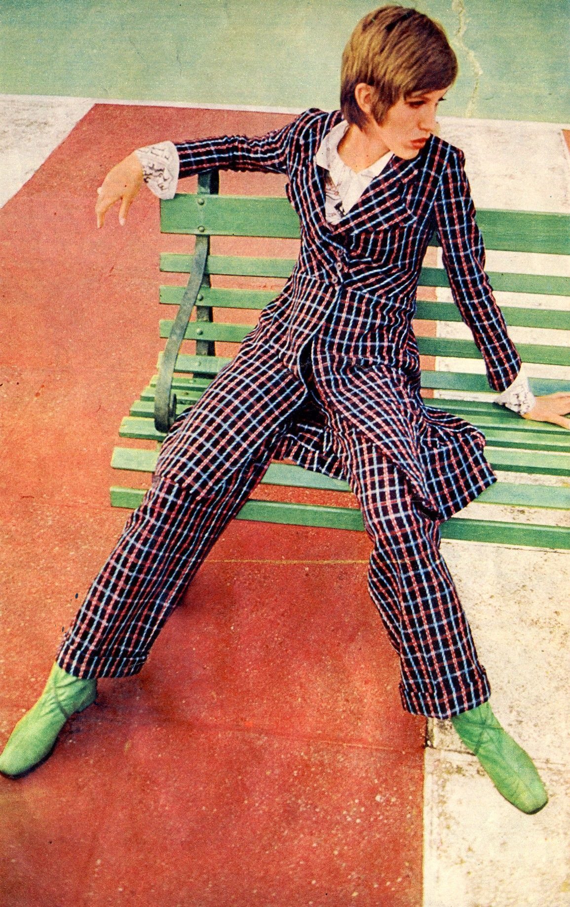 Person in a colorful plaid suit and green shoes sitting on a green bench.