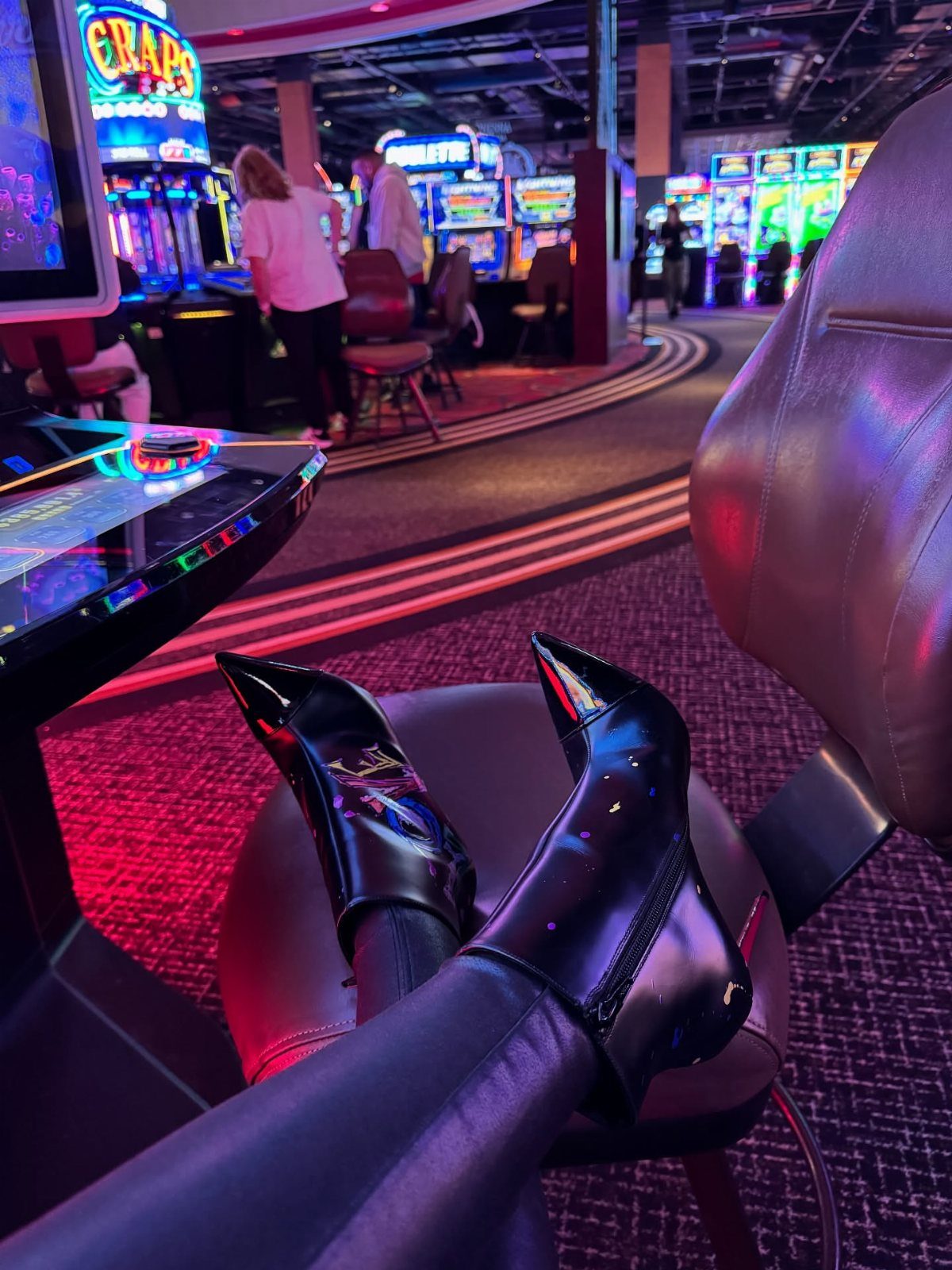 Shiny black pointed-toe ankle boots worn by a person sitting in a casino.