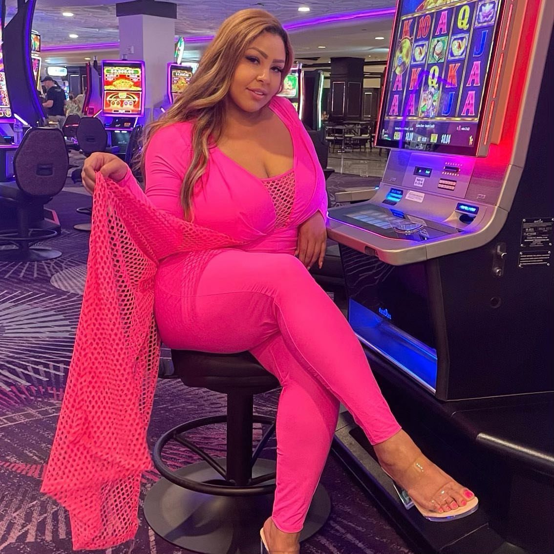 Woman in a bright pink jumpsuit sitting on a stool in a casino setting.