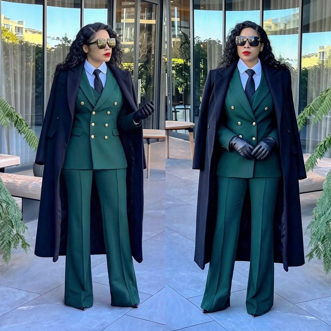 Woman in a dark green double-breasted pantsuit and long black coat standing outdoors.
