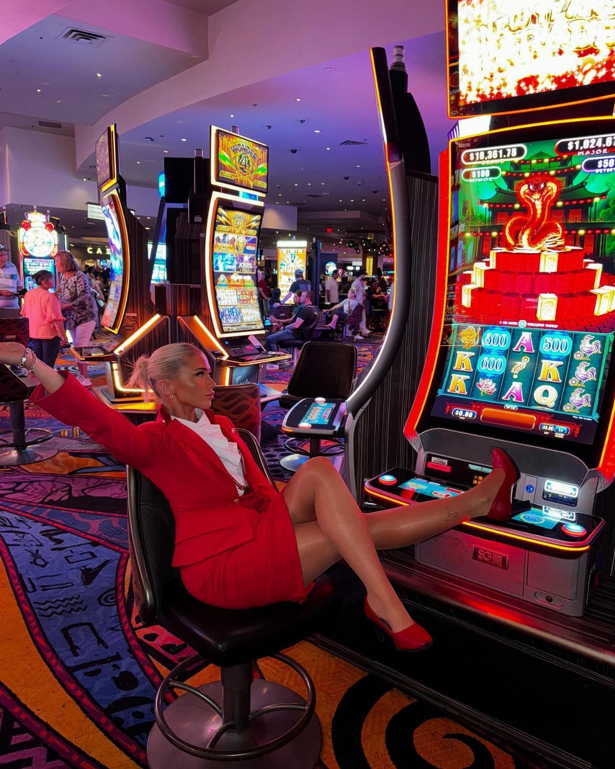 Woman in a red blazer and skirt posing while sitting at a casino slot machine.